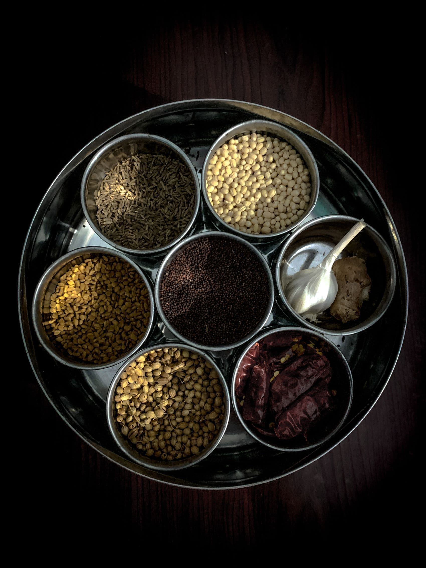 A steel masala dabba holding cumin, mustard seed, coriander, dal and dried chillies